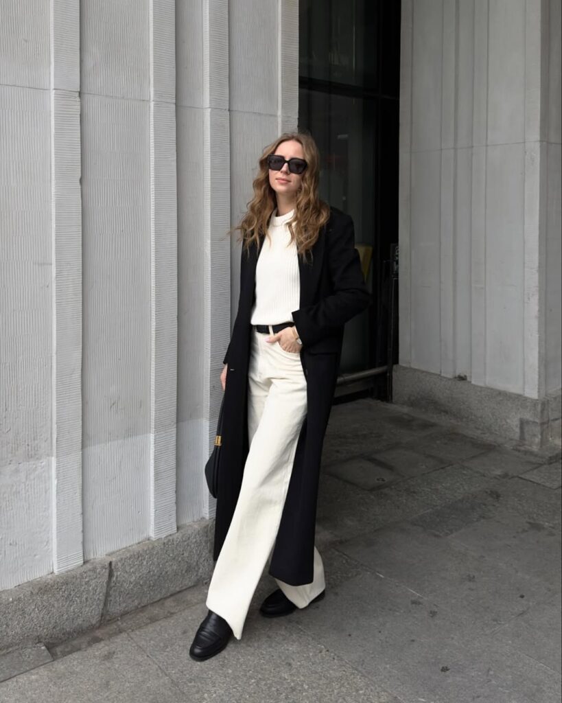 woman in long black coat and white trousers standing against white textured wall