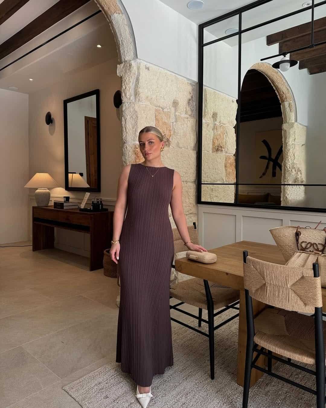 woman wearing a sleeveless brown pleated maxi dress standing in a modern neutral toned interior