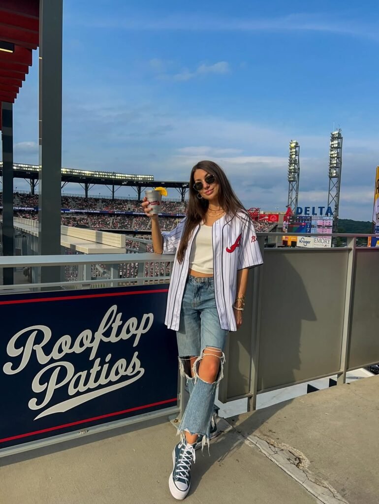 Woman in pinstripe Braves jersey and ripped jeans holding drink at stadium patio