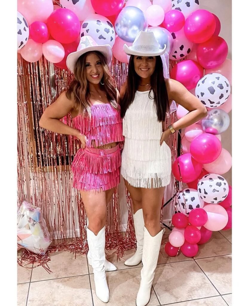 two women in pink and white fringe outfits posing against balloon backdrop