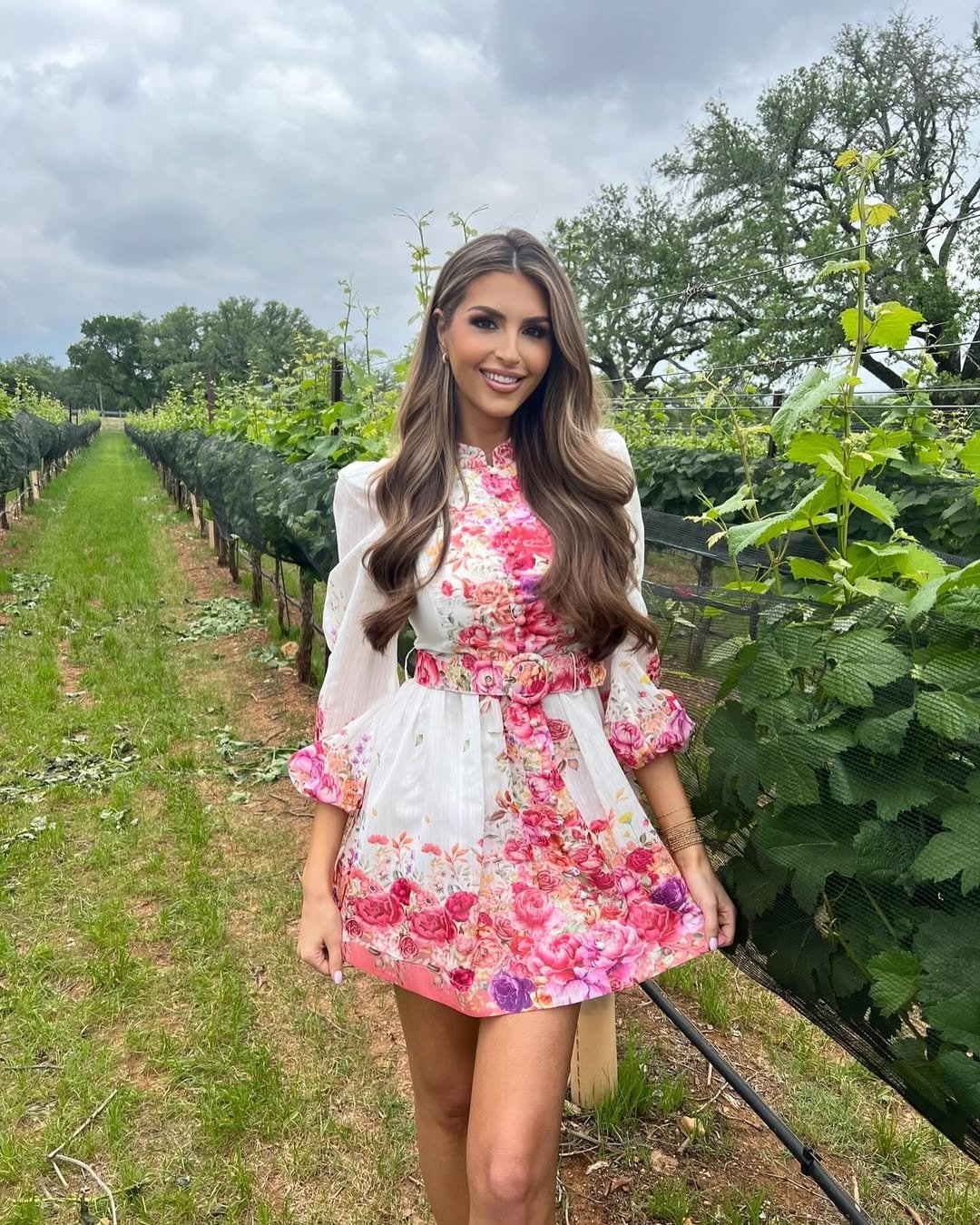 smiling woman wearing a floral mini dress standing in a lush green vineyard