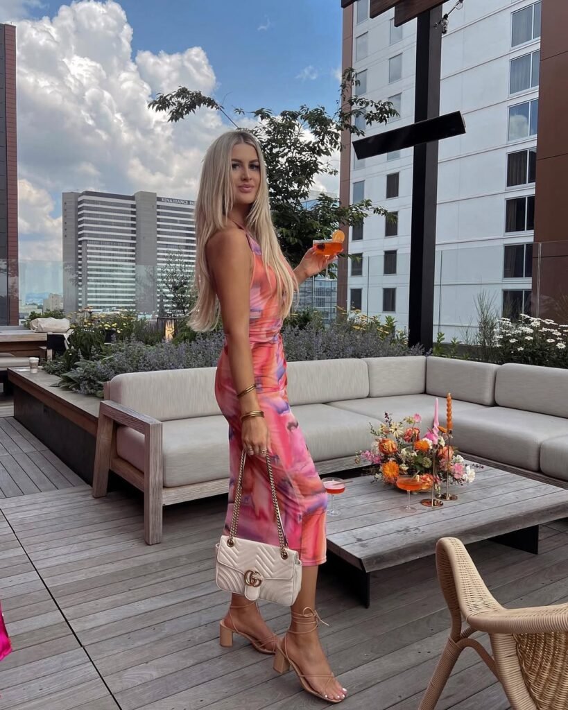 woman in pink floral midi dress holding cocktail on modern rooftop lounge deck