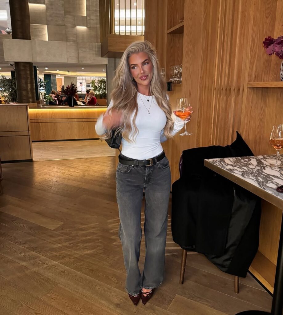 woman in a white top and grey jeans holding a drink in a restaurant
