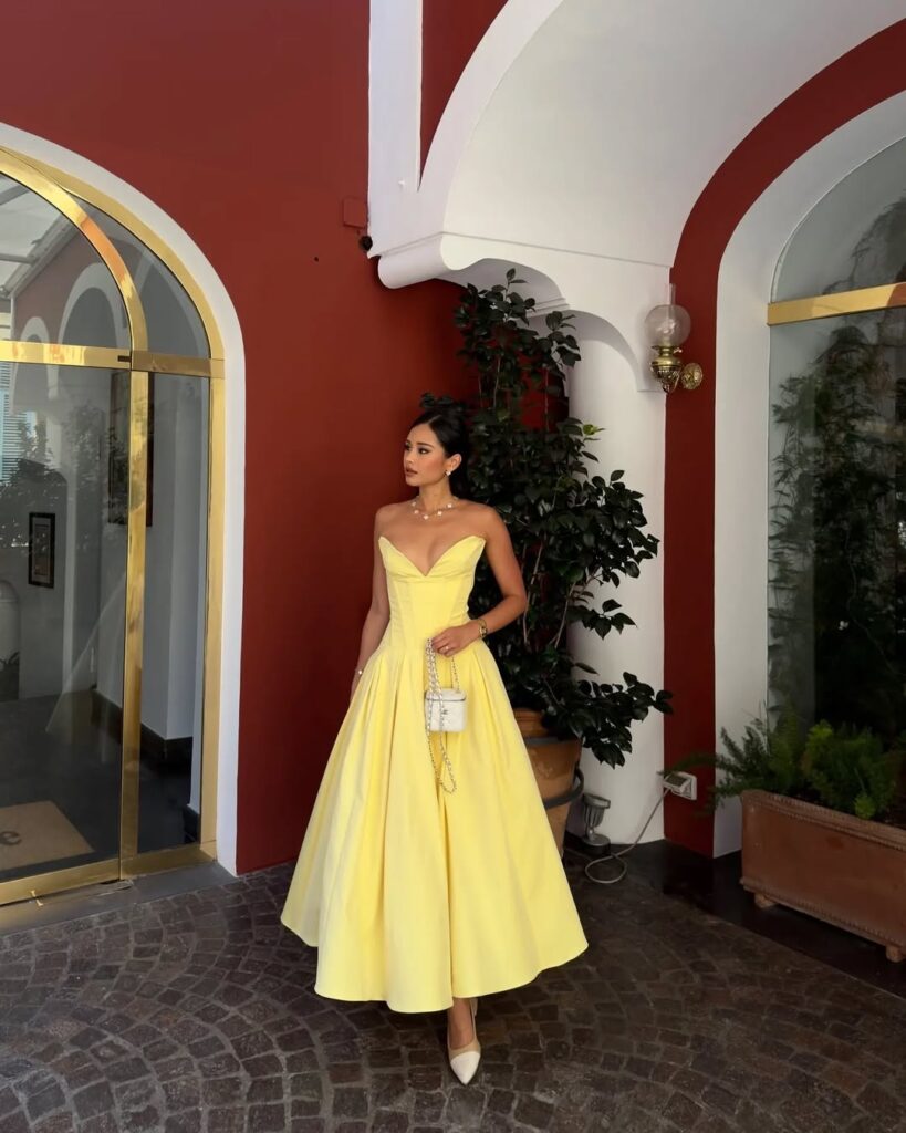 woman in strapless yellow gown holding white handbag standing near red arched walls