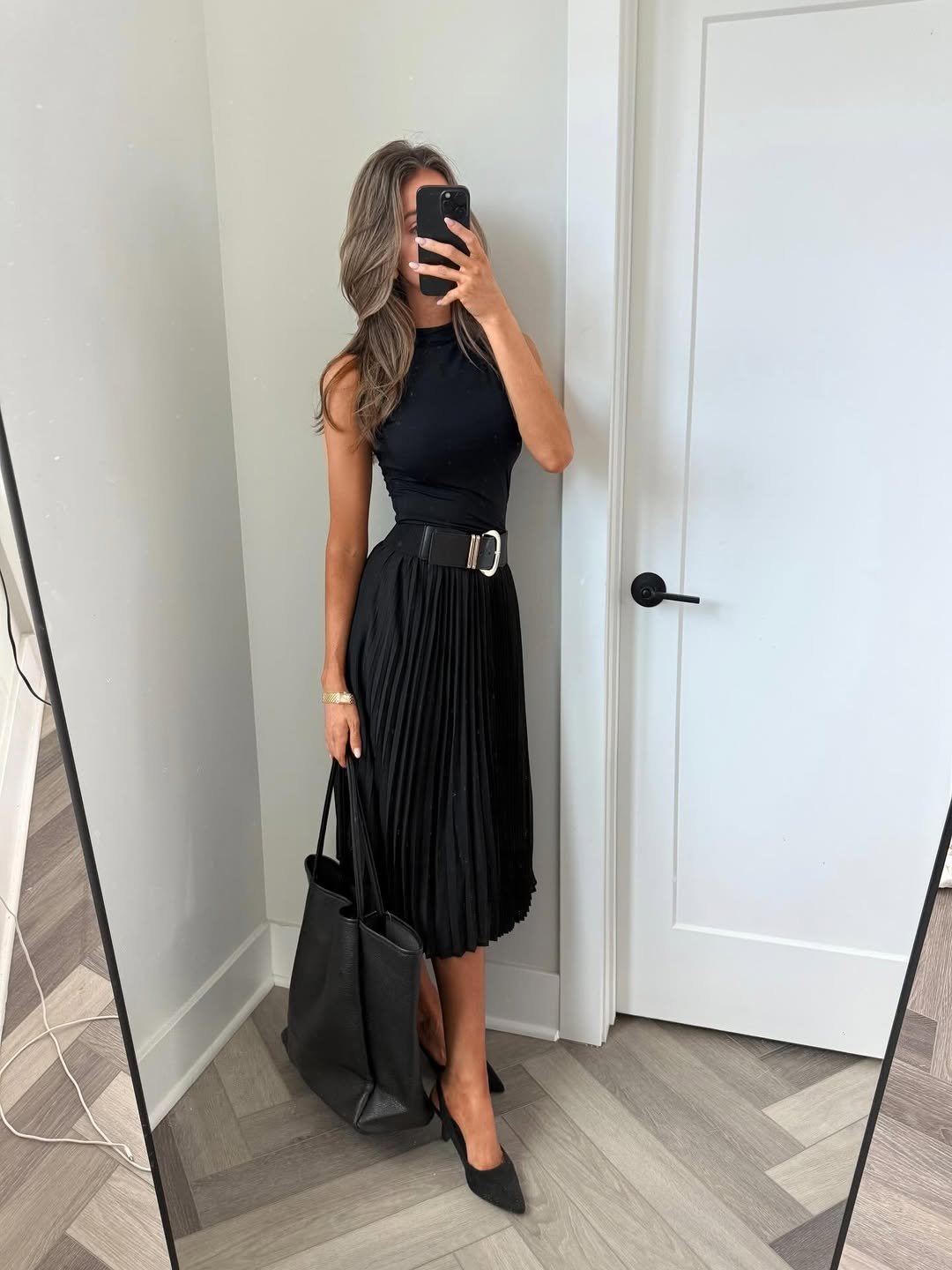 woman taking a mirror selfie wearing a black sleeveless top and pleated midi skirt