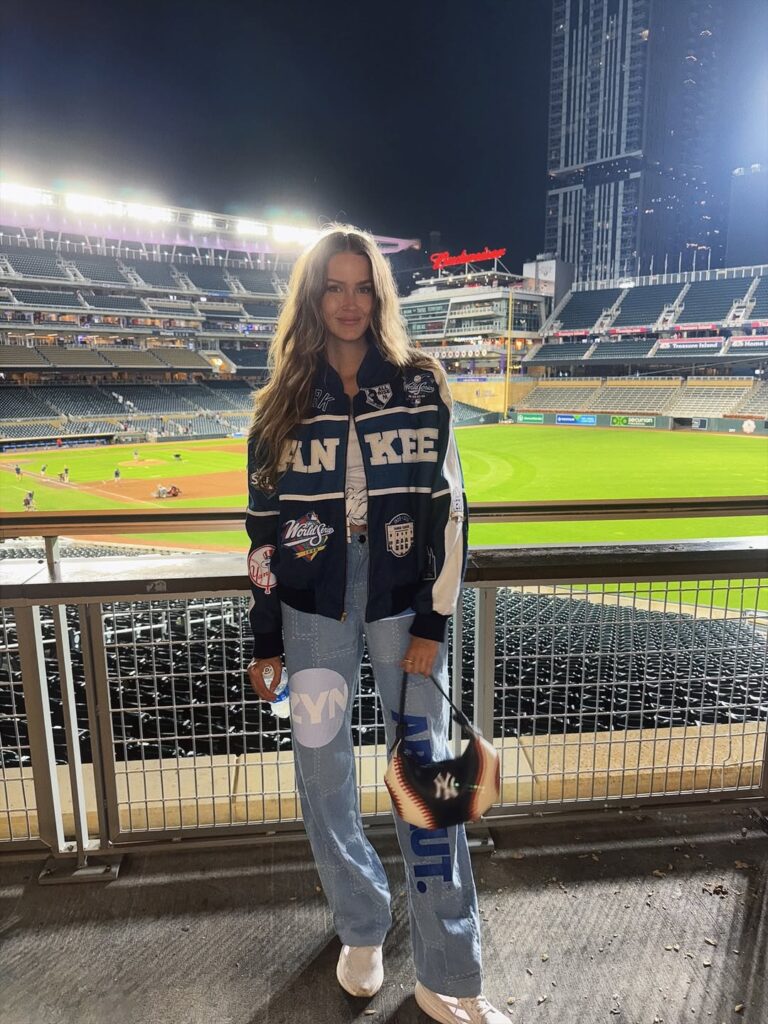woman in vintage yankees jacket and graphic jeans standing in front of stadium field