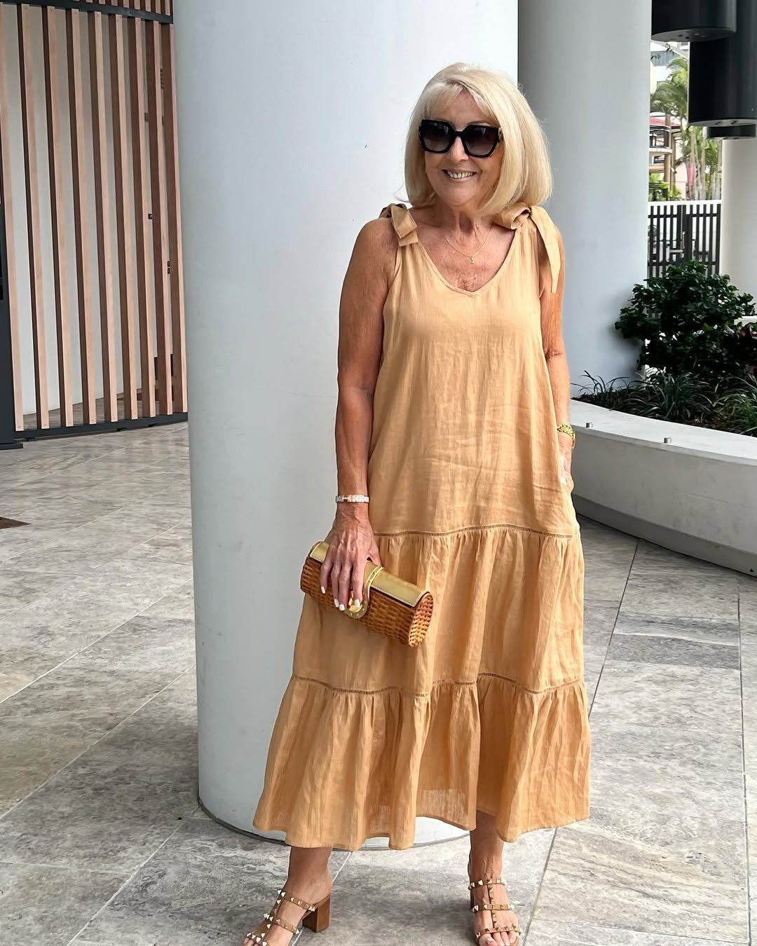 woman wearing a sleeveless tan linen tiered maxi dress holding a small woven clutch bag