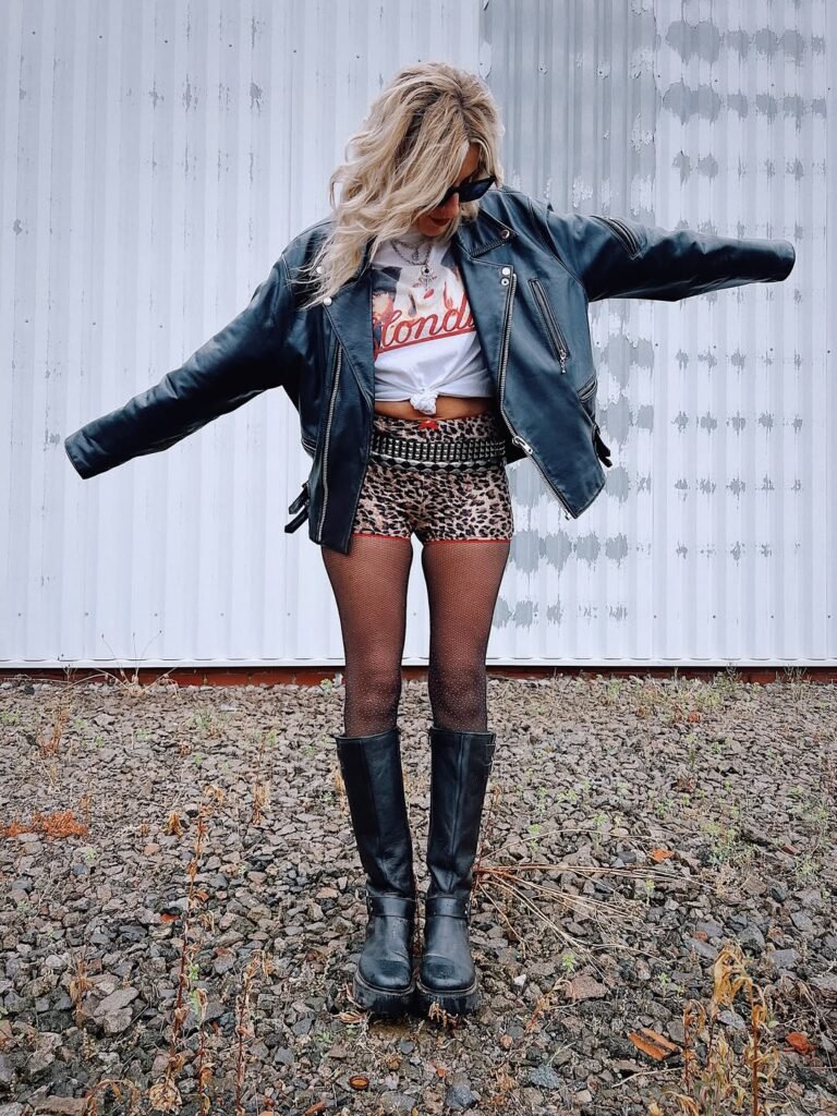 blonde woman in leather jacket and leopard print shorts standing on gravel ground