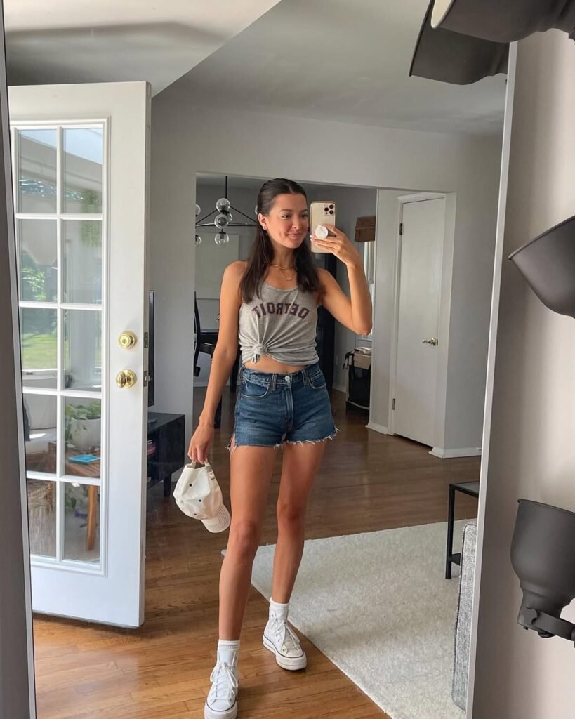 Mirror selfie of woman in Detroit tank top and denim shorts holding white cap