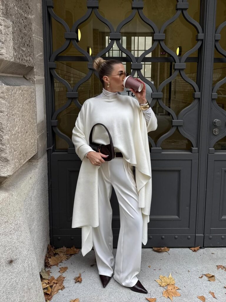 woman in white cape and trousers drinking coffee in front of dark metal gate