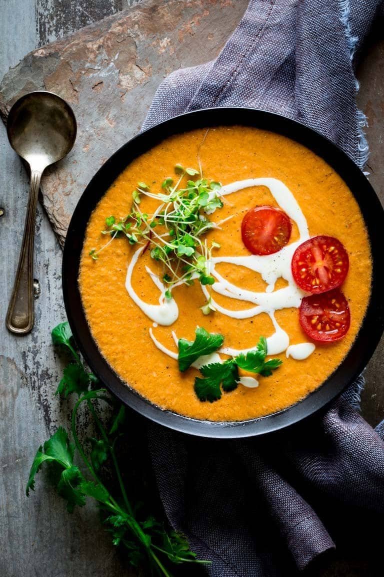 orange gazpacho soup topped with cream, microgreens, and sliced tomatoes in a black bowl.