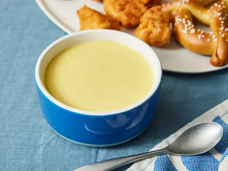 creamy yellow honey mustard dipping sauce in a blue bowl with pretzels and chicken nuggets.