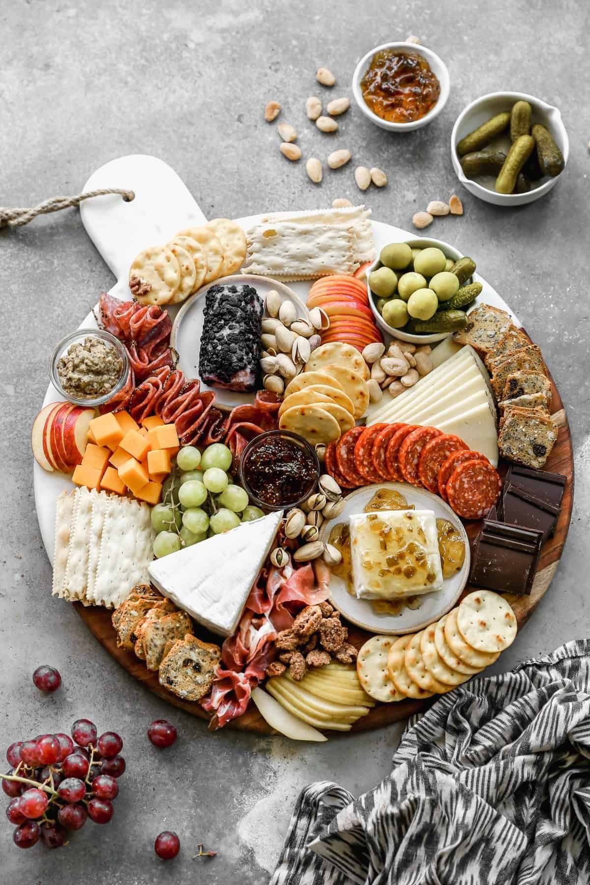 large round charcuterie board with various cheeses, meats, crackers, fruits, and nuts on gray stone.
