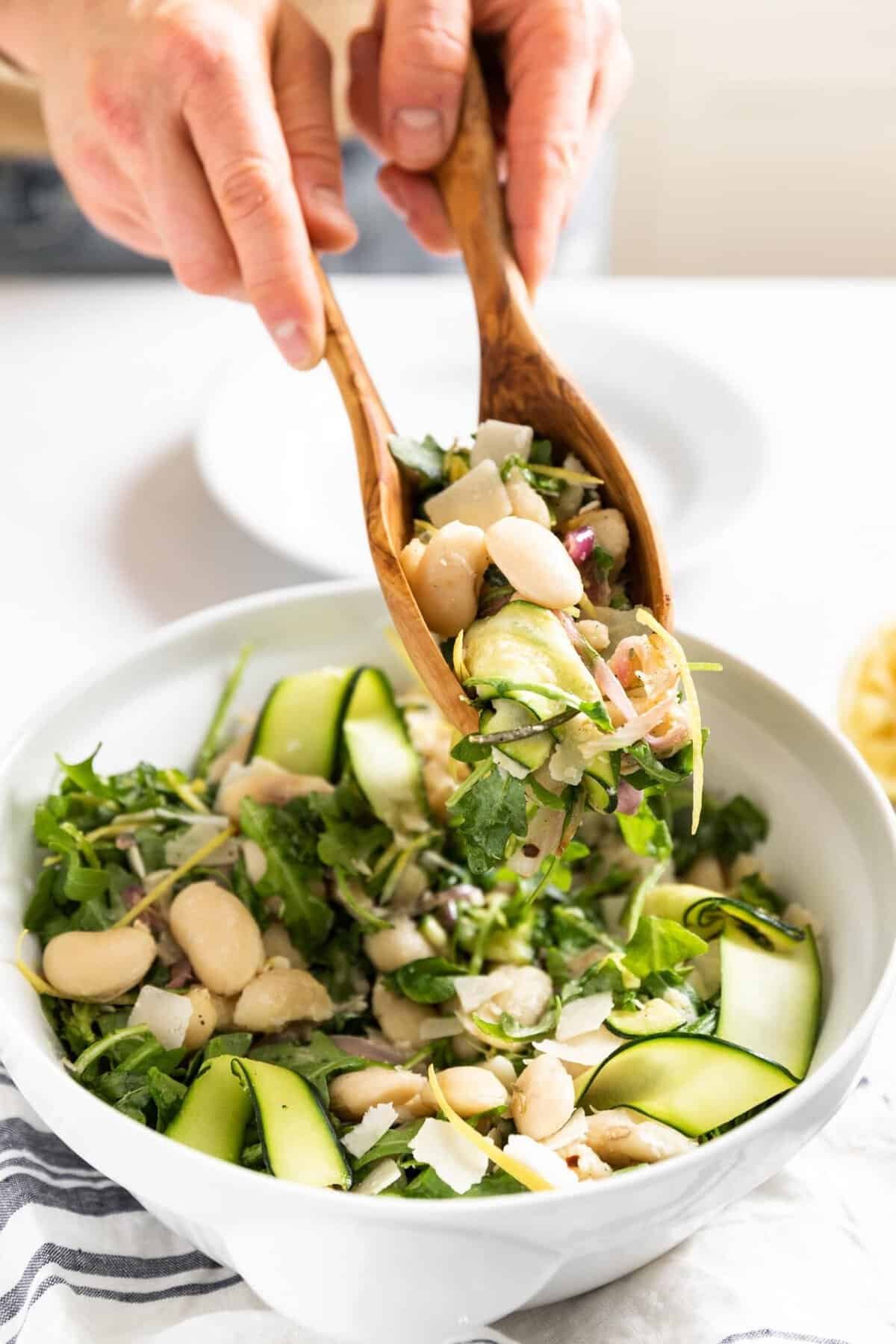 white bean salad with shaved zucchini and arugula being served with large wooden spoons.