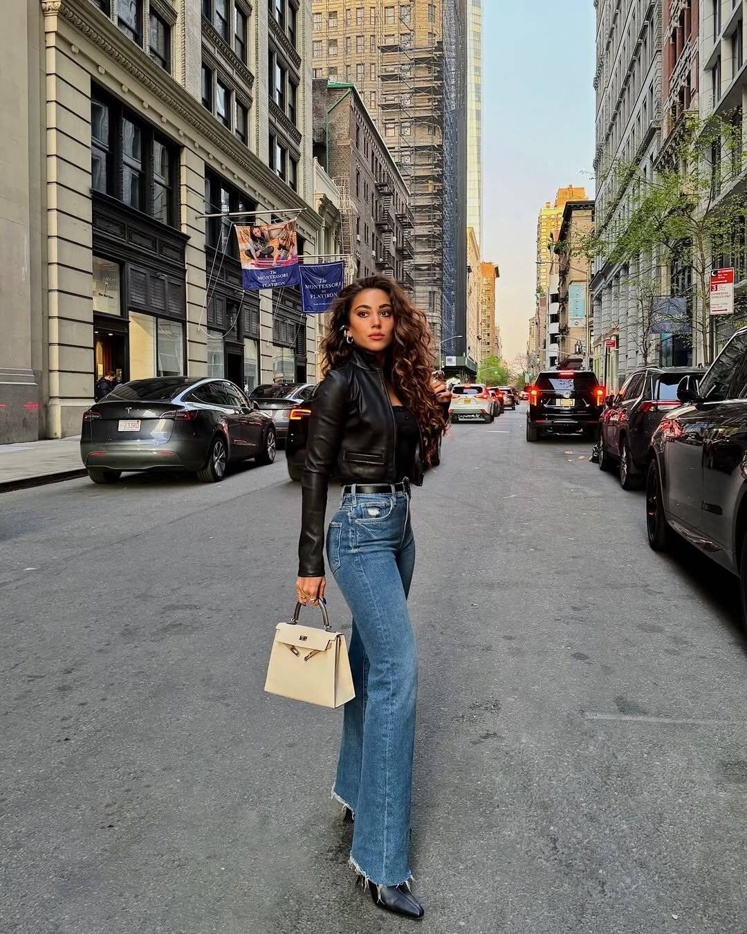 woman in black leather jacket and flared jeans holding a beige handbag in new york city