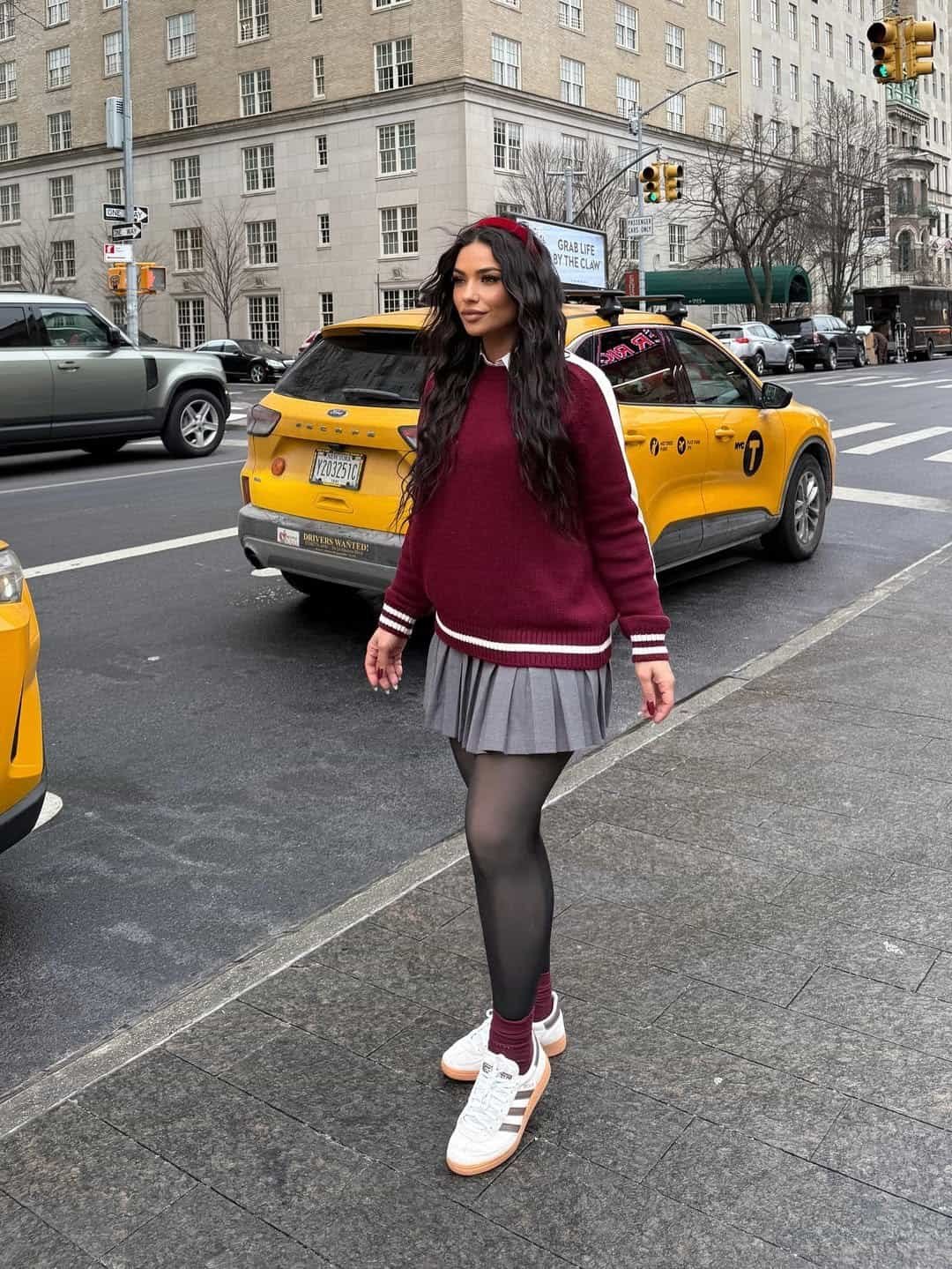woman wearing maroon sweater and grey pleated skirt walking past a yellow new york taxi