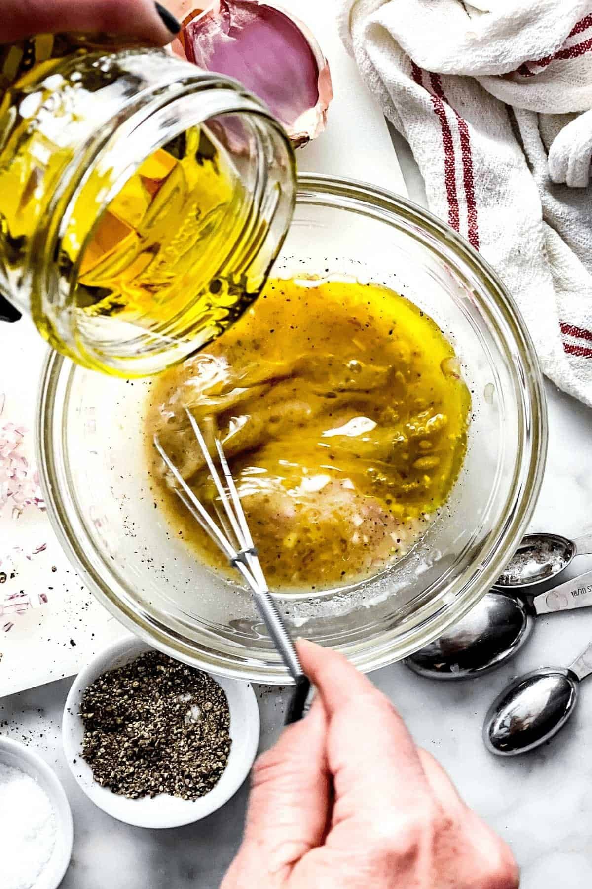 olive oil pouring into a glass bowl with a whisk and vinaigrette ingredients.