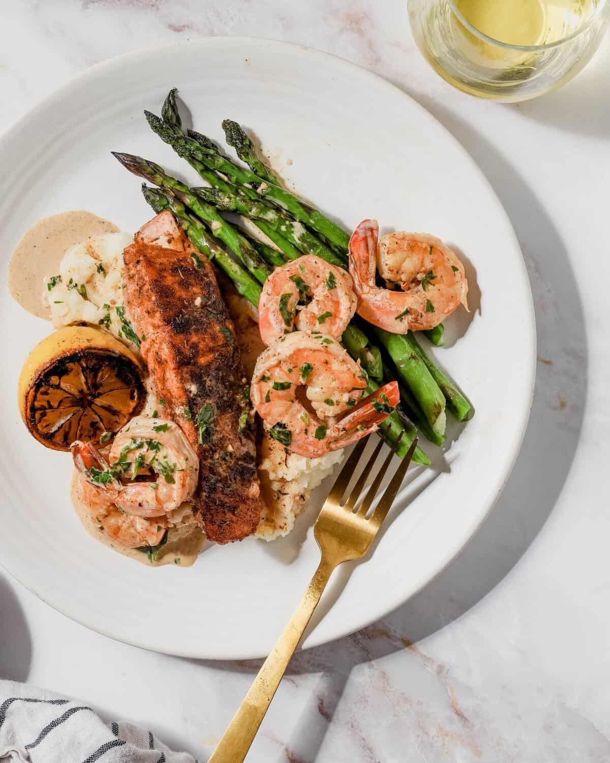 blackened salmon fillet served with seared shrimp, asparagus, and mashed potatoes on a white plate.