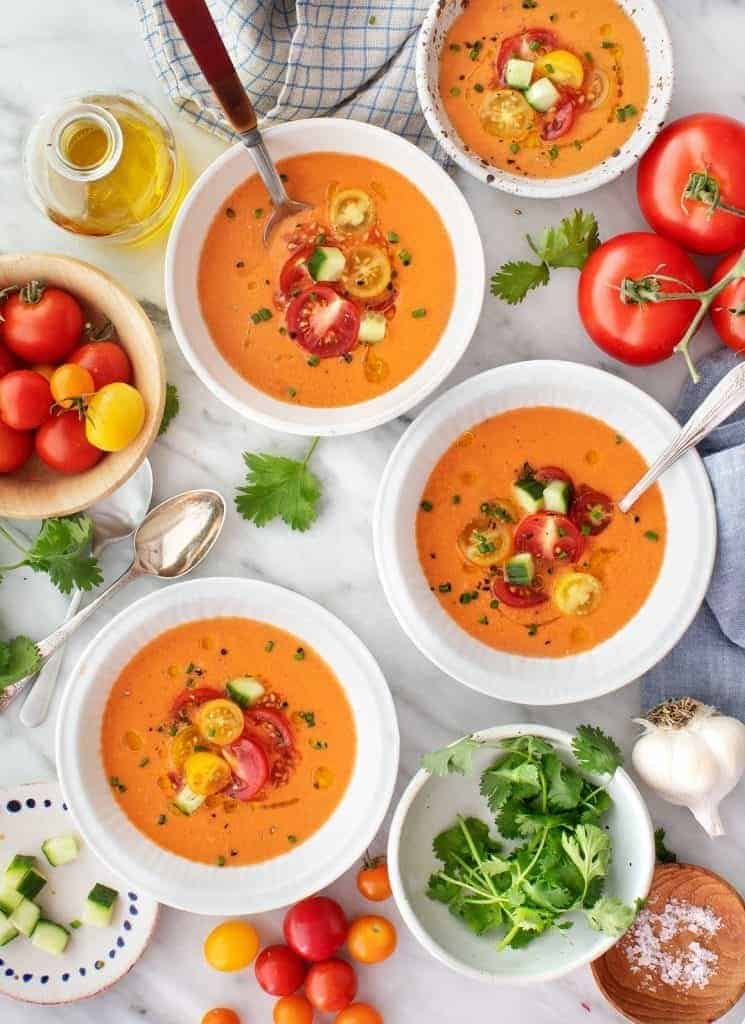 four bowls of chilled tomato gazpacho garnished with diced cucumbers and colorful cherry tomatoes.