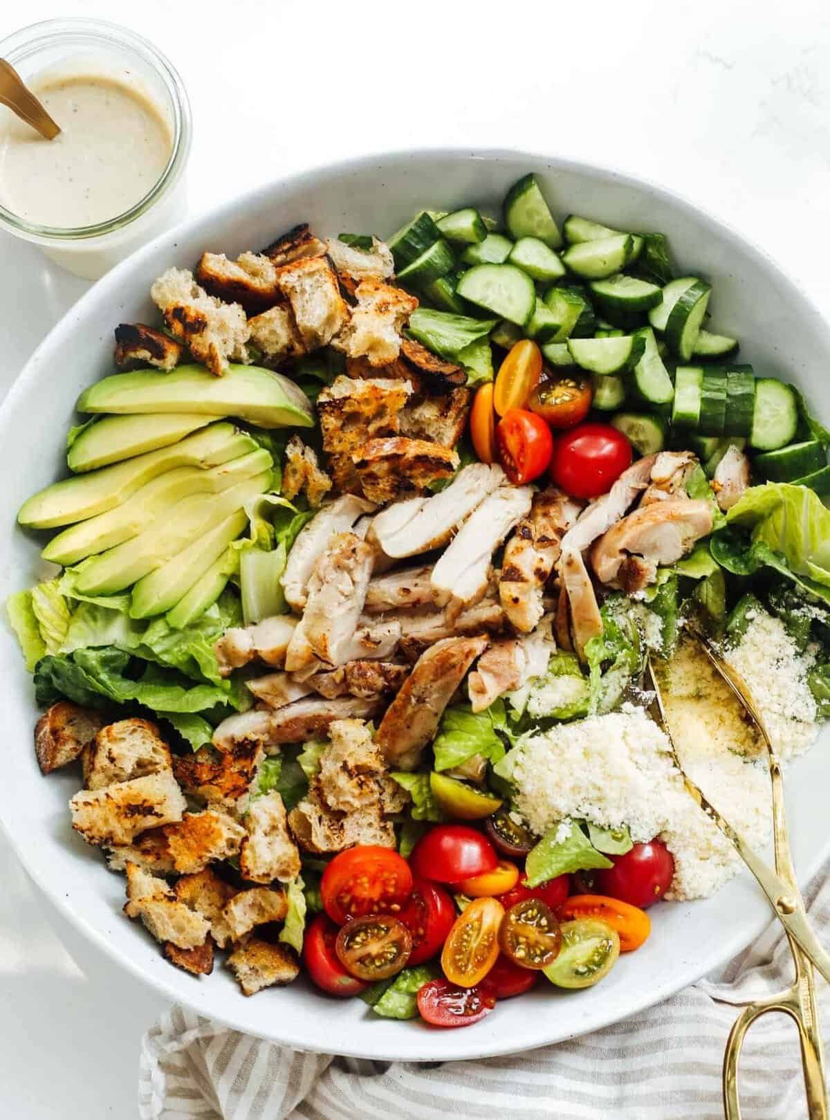 grilled chicken caesar salad with avocado, tomatoes, cucumbers, and croutons in a bowl.