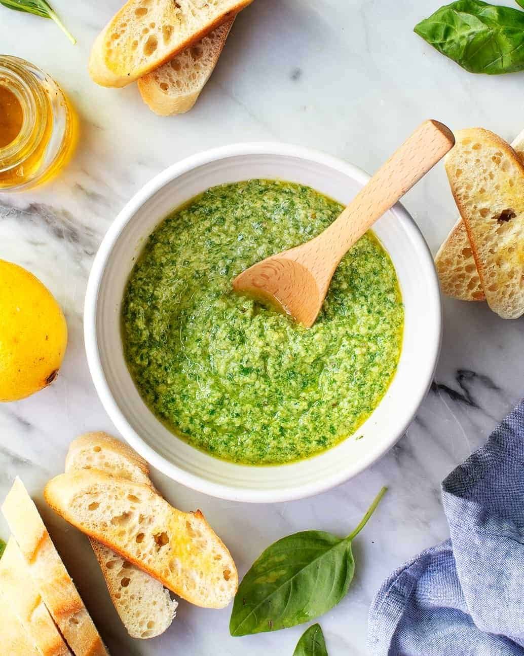 fresh basil pesto in a white bowl with a wooden spoon and baguette slices.
