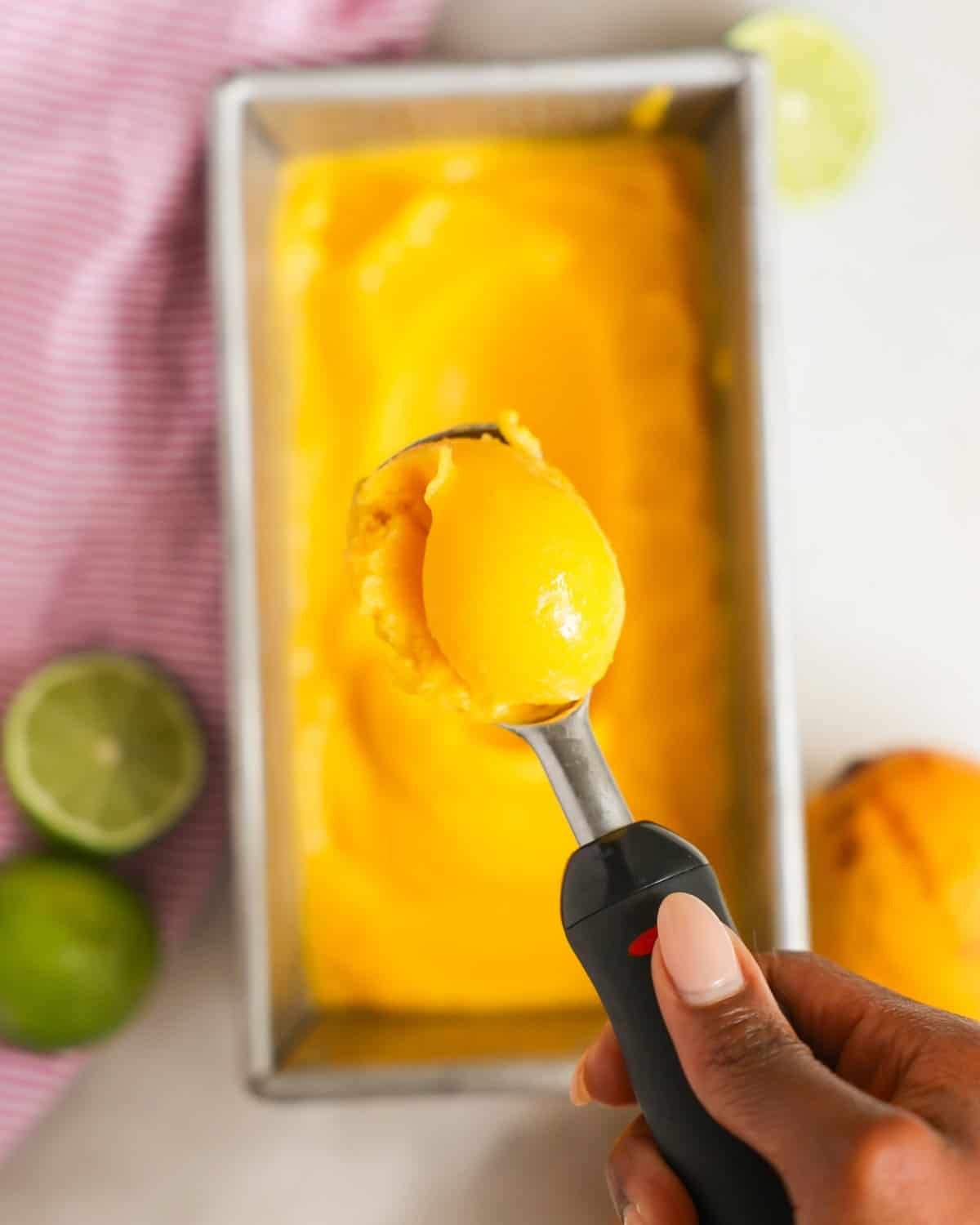 a hand holding a metal scoop of bright yellow mango sorbet over a metal container