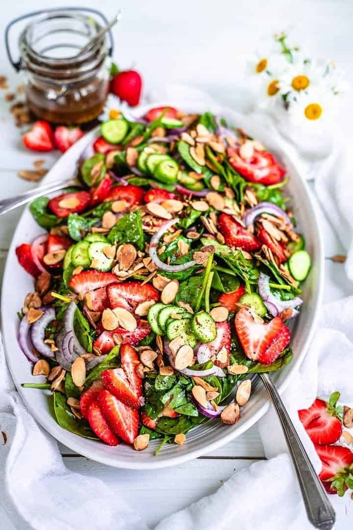 strawberry spinach salad with sliced cucumbers, red onions, and almonds on a white platter.