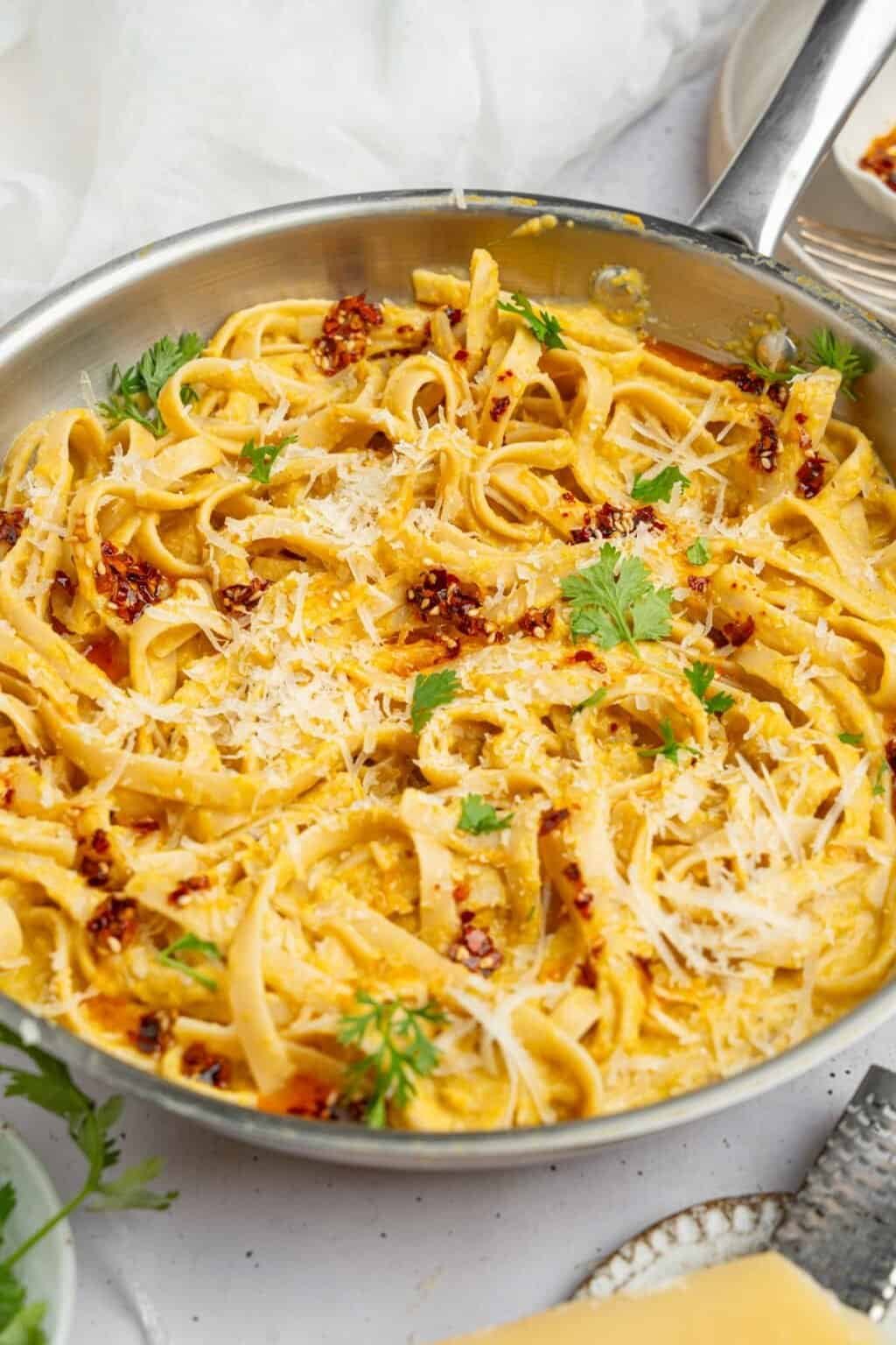 creamy fettuccine pasta in a stainless steel pan topped with parmesan, chili flakes, and parsley.