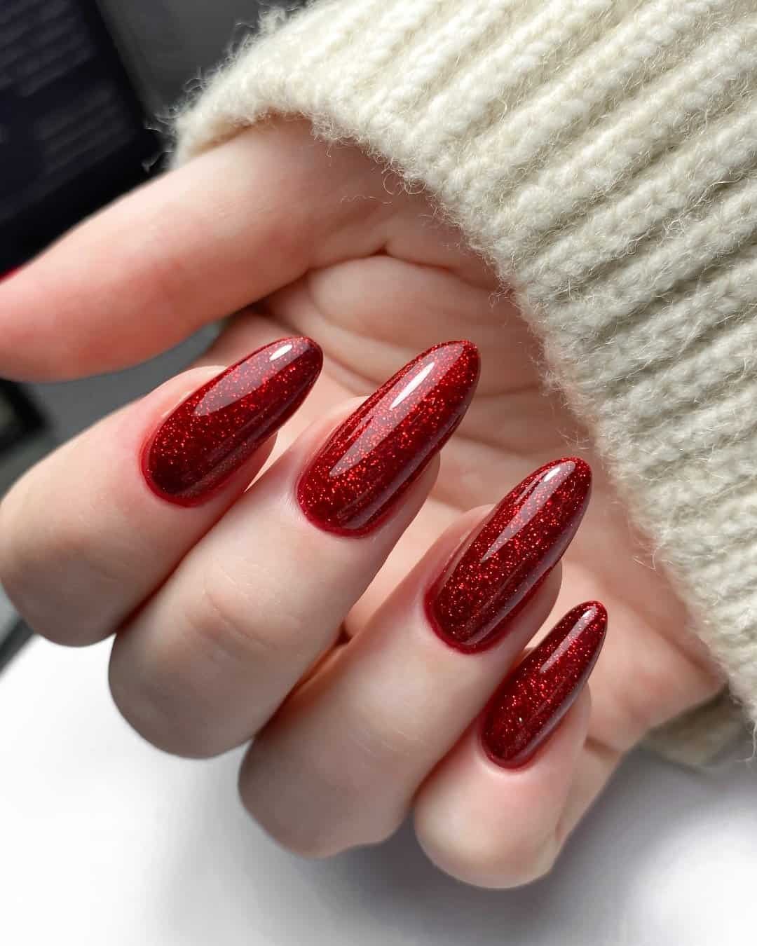 long almond nails with a shimmering ruby red glitter finish and sweater cuff.