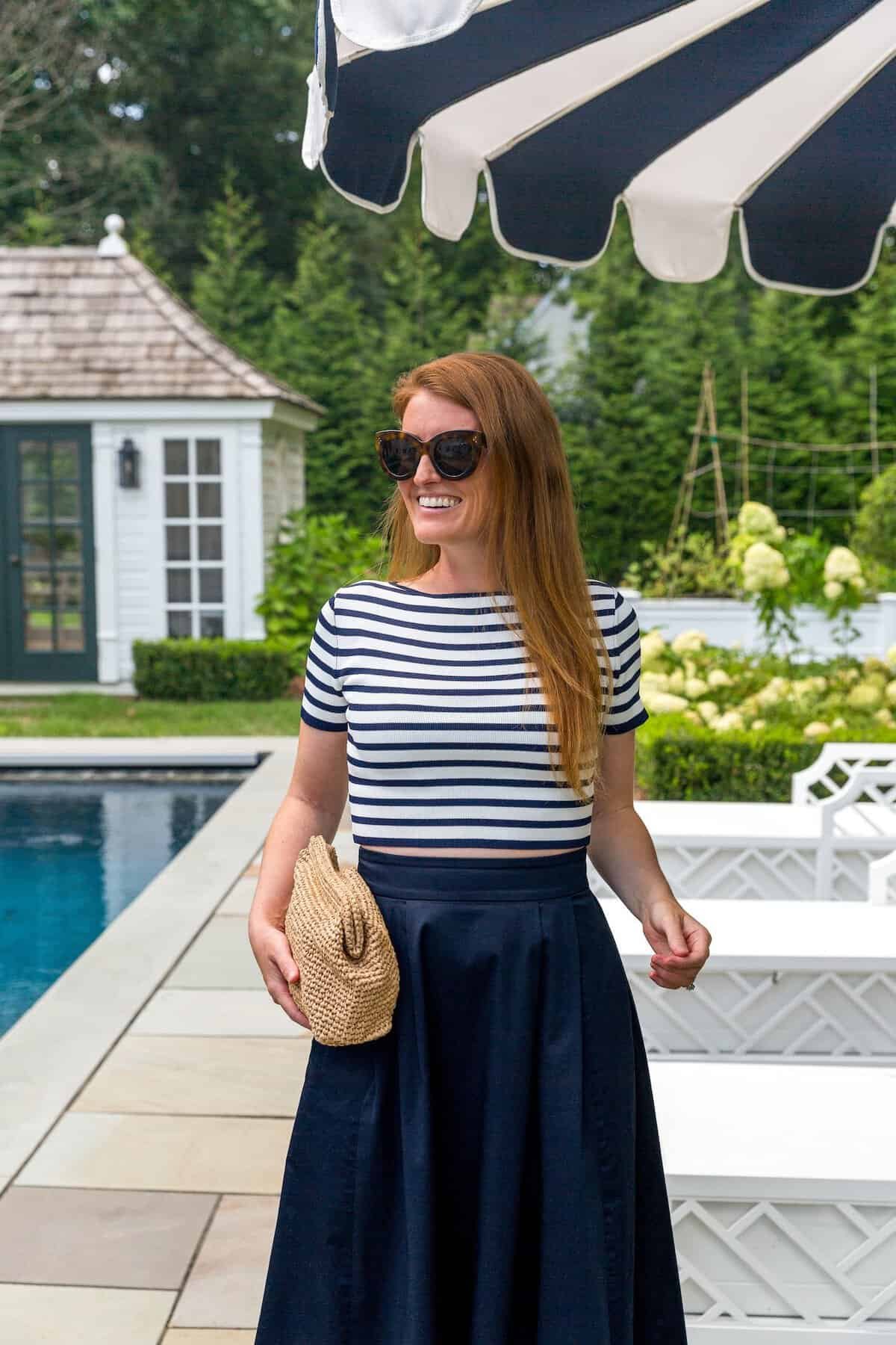 woman in striped top and navy skirt holding straw clutch by pool.