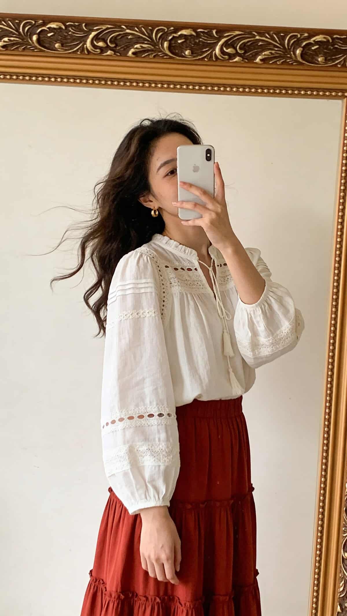 woman in white bohemian blouse and red tiered skirt taking a mirror selfie.