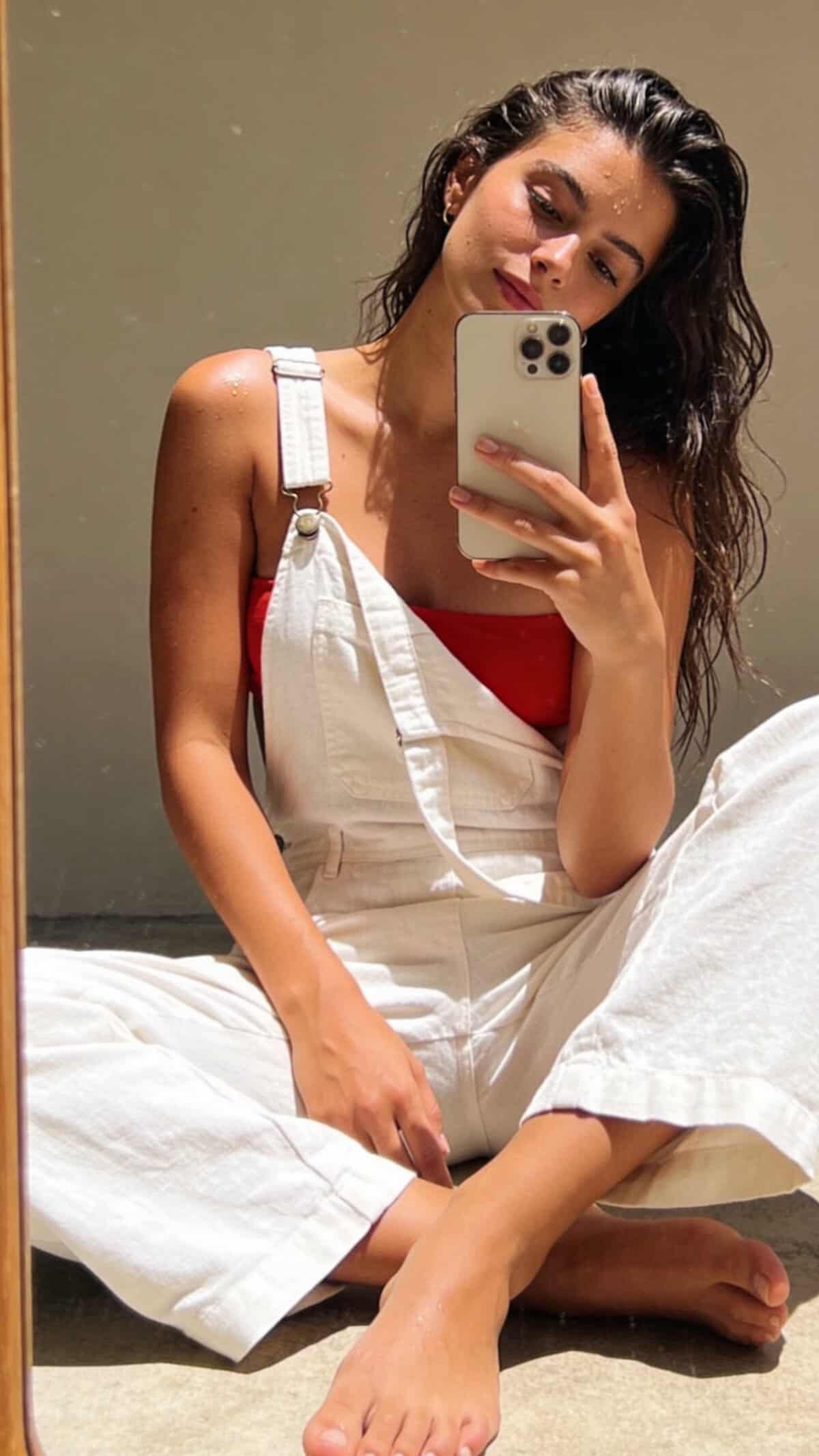 woman sitting on floor in white overalls and red top taking mirror selfie.