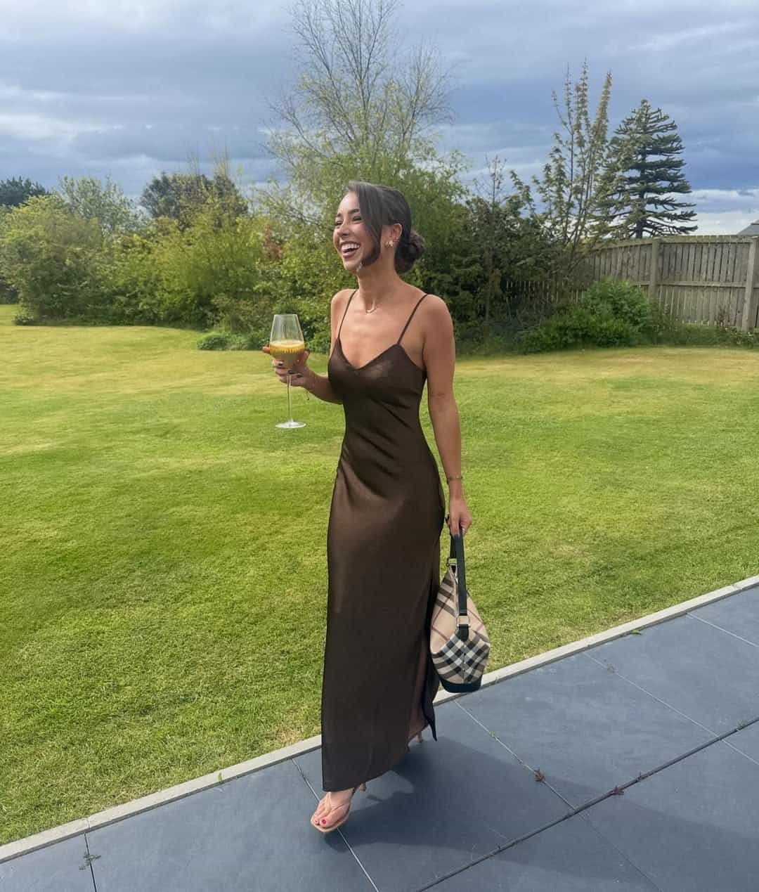 smiling woman in a dark brown satin slip dress holding a wine glass outdoors