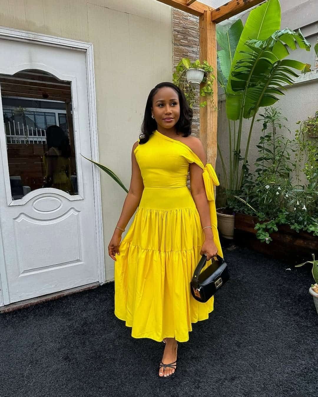 woman wearing a bright yellow one shoulder tiered midi dress holding a black handbag