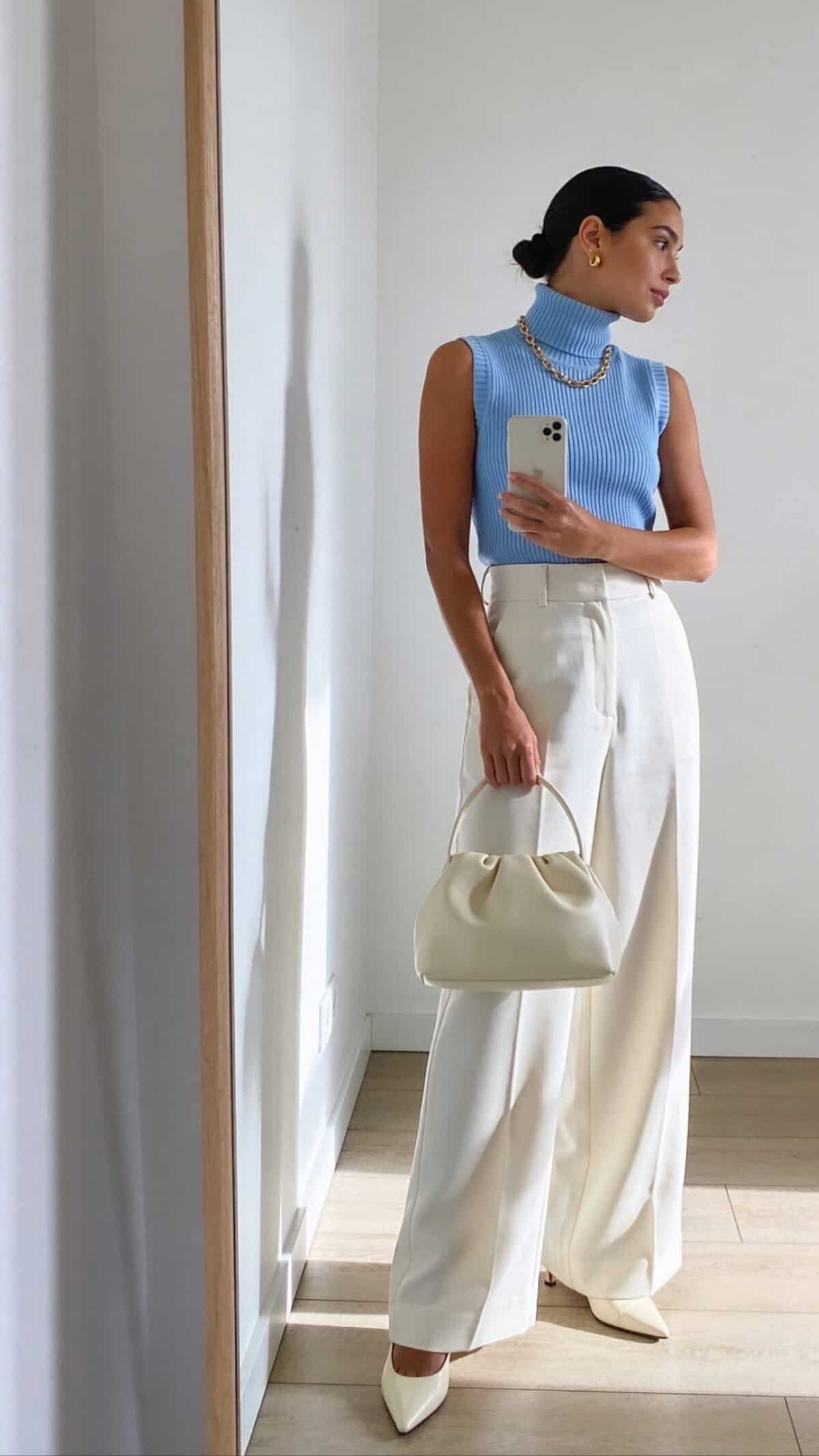 woman in light blue sleeveless turtleneck and cream wide leg trousers holding a handbag
