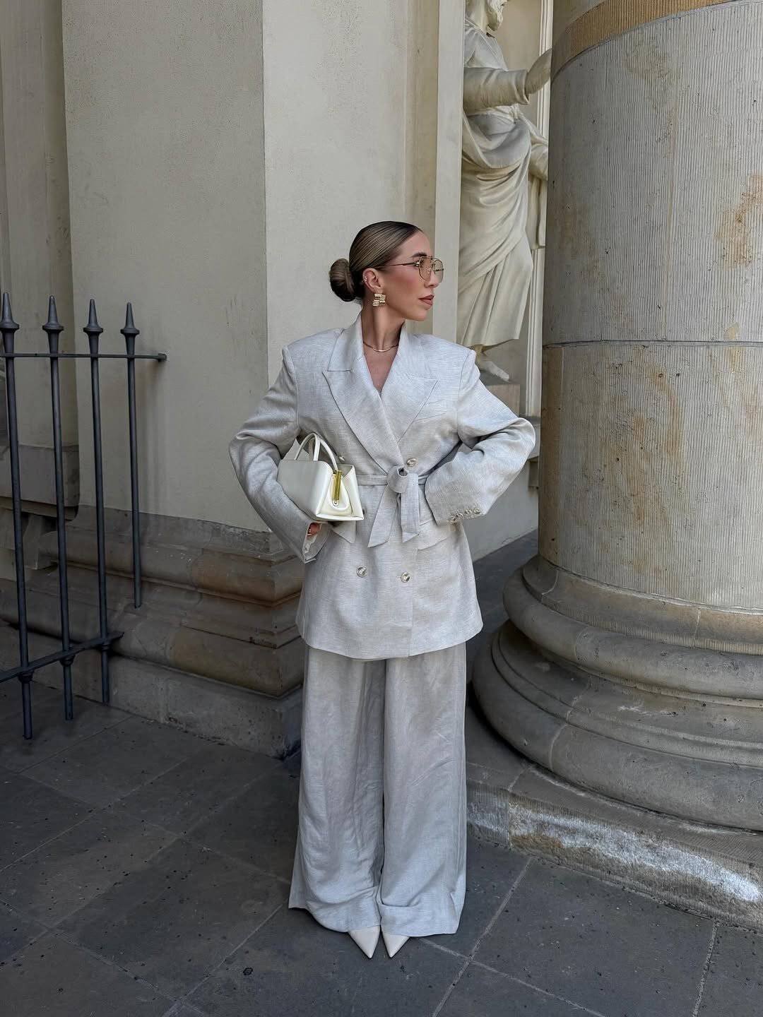 woman wearing oversized cream linen pantsuit and carrying a small white handbag outdoors