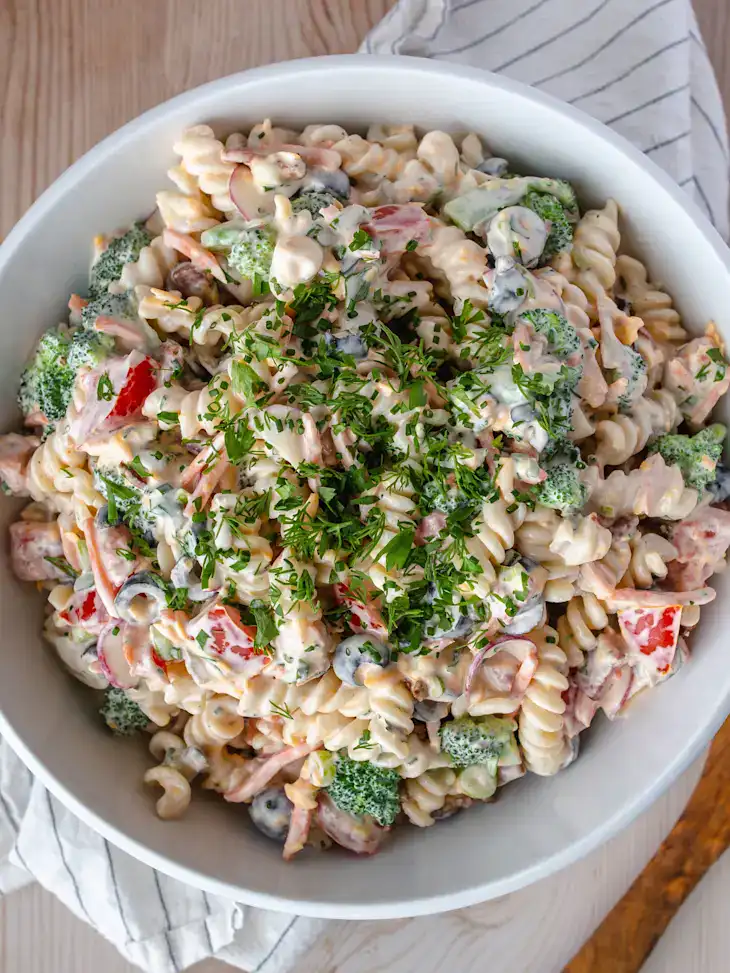 creamy rotini pasta salad with broccoli, olives, and red peppers topped with fresh herbs