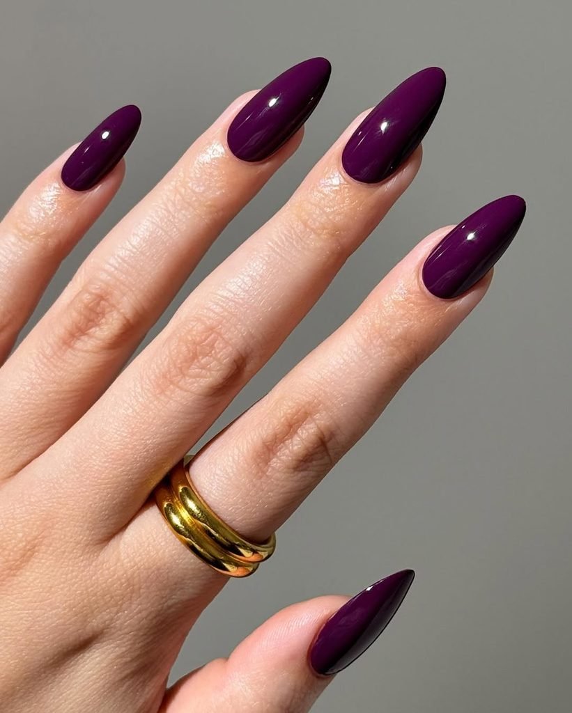 deep plum purple glossy almond nails on a hand wearing a gold ring