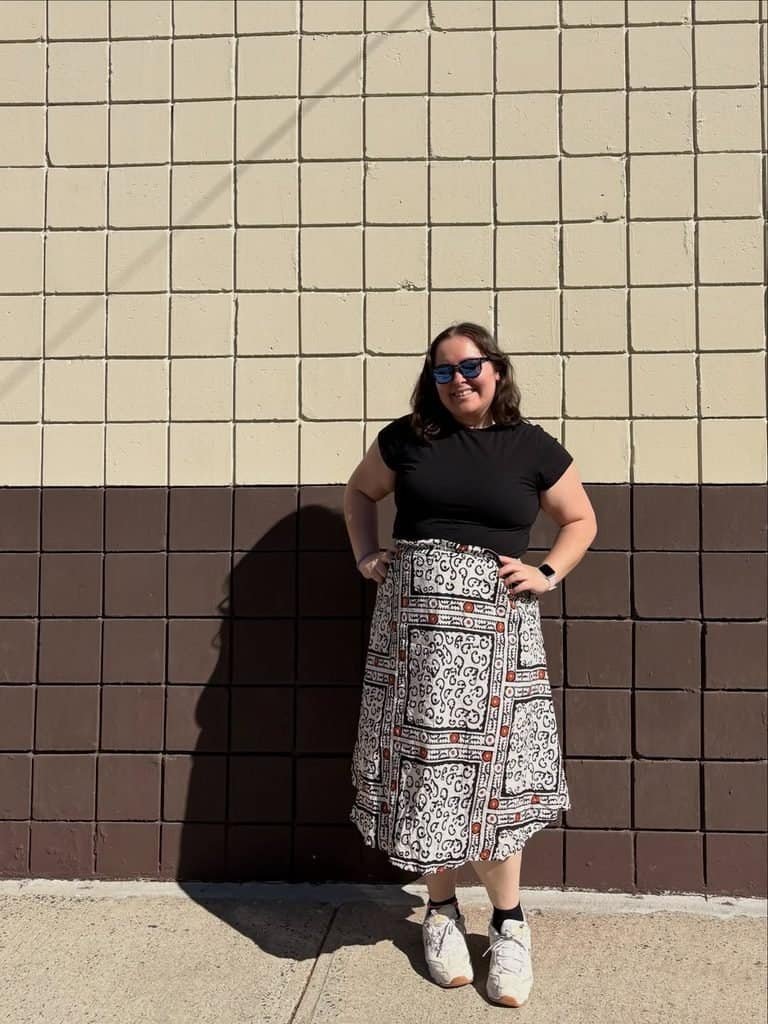 woman standing in front of a tiled wall wearing a black t-shirt and pattern skirt