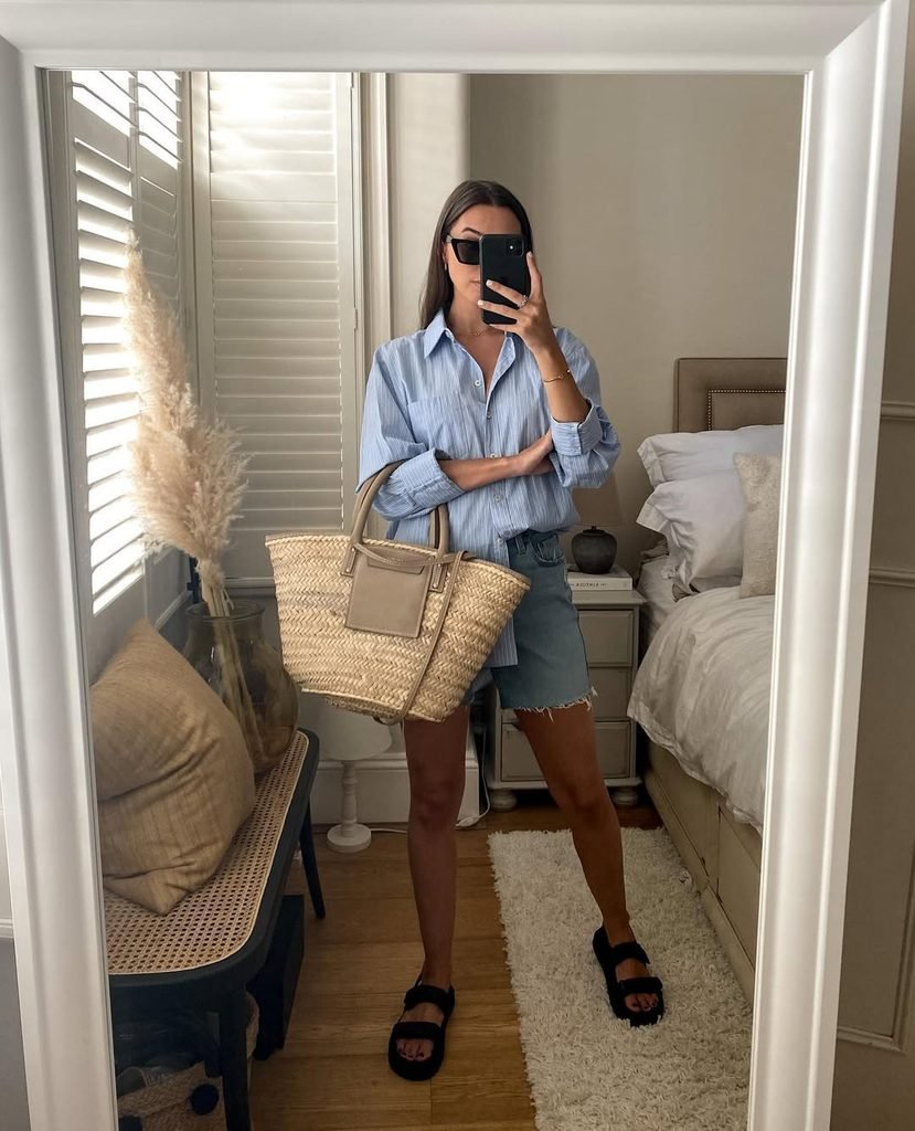 woman wearing blue striped button-down shirt denim shorts and holding woven tote bag mirror selfie