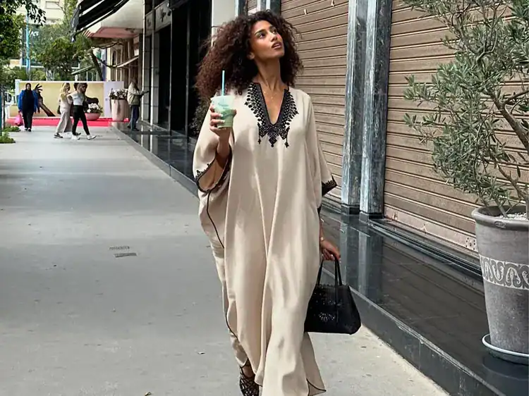 woman in a beige kaftan walking on a city sidewalk holding a drink