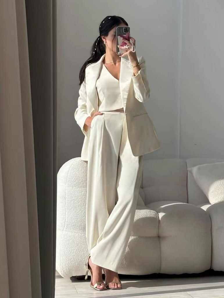 woman standing by white couch wearing monochromatic cream three-piece pantsuit and gold heels