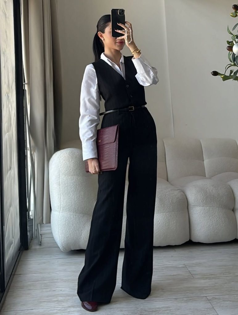 woman wearing black vest, matching black trousers, white shirt, holding burgundy laptop sleeve