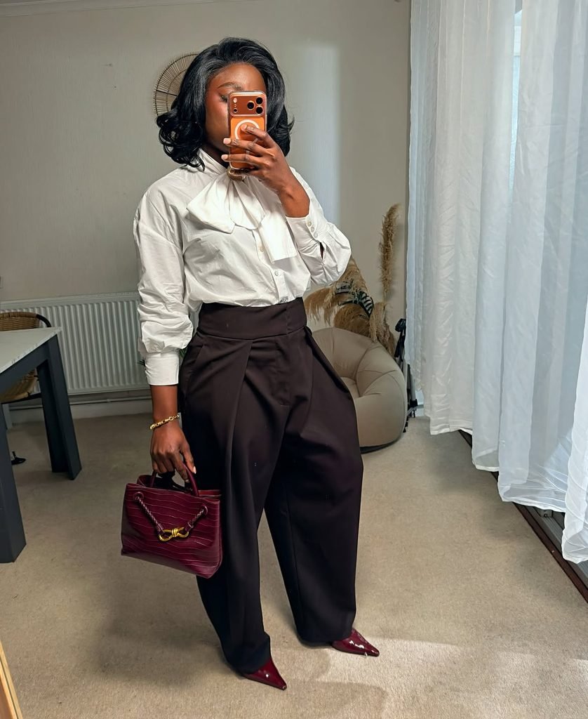 woman wearing white bow blouse, high-waisted dark brown trousers, holding burgundy textured purse