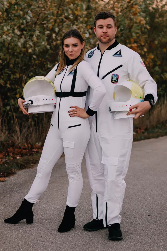 man and woman wearing matching white astronaut costumes while holding space helmets outdoors