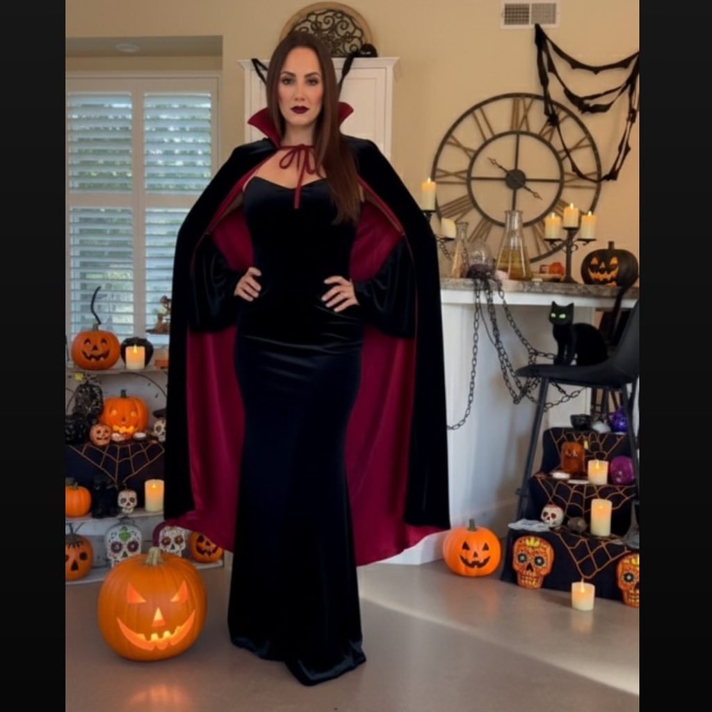 woman dressed as a vampire wearing a black gown and red cape near pumpkins