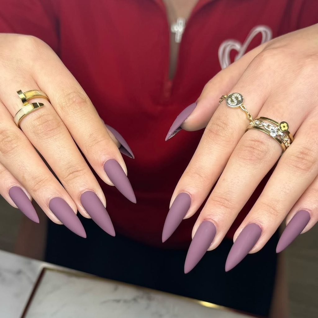 long stiletto nails with a smooth matte purple finish wearing gold rings