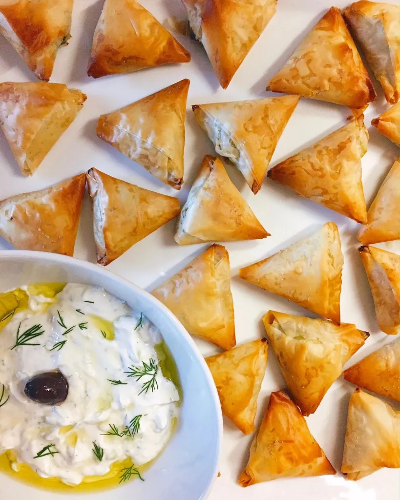 golden brown triangle pastries arranged on a tray next to a bowl of tzatziki dip