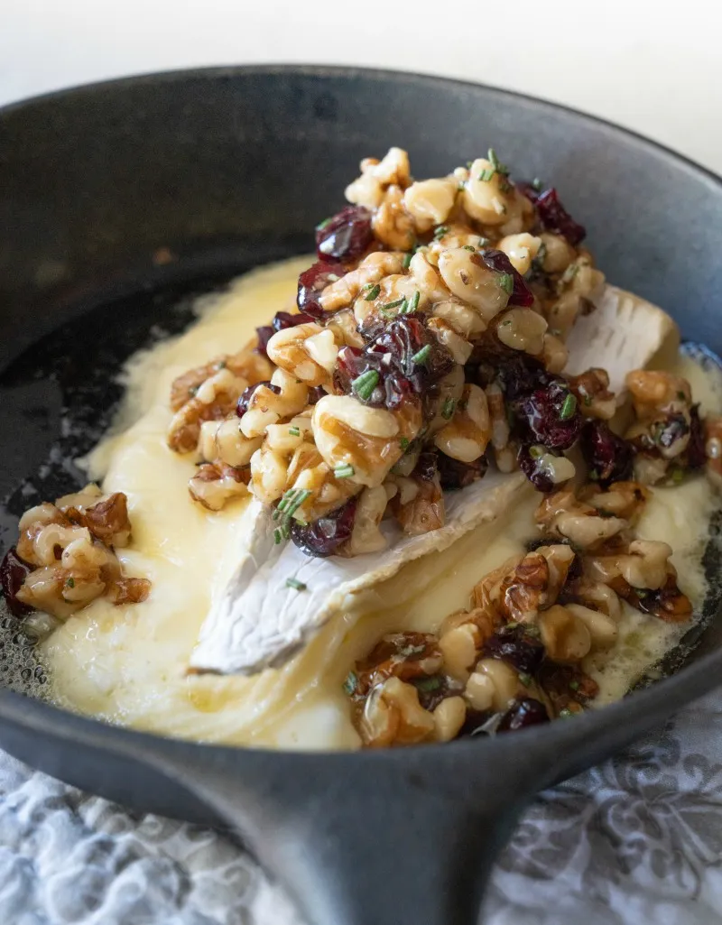 baked brie cheese in a skillet topped with chopped walnuts and dried cranberries