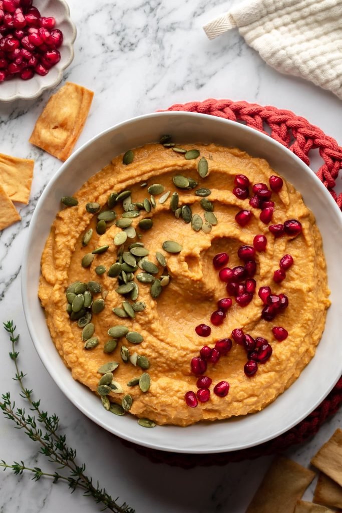 pumpkin hummus dip topped with pumpkin seeds and pomegranate seeds in a white bowl